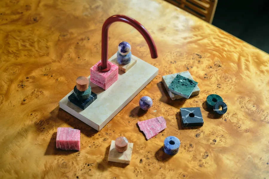 Grifo de Dornbracht Atelier diseñado por Fabian Freytag con caño rojo curvo, cubo de rodocrosita y manijas de piedras apiladas sobre una mesa de madera de raíz