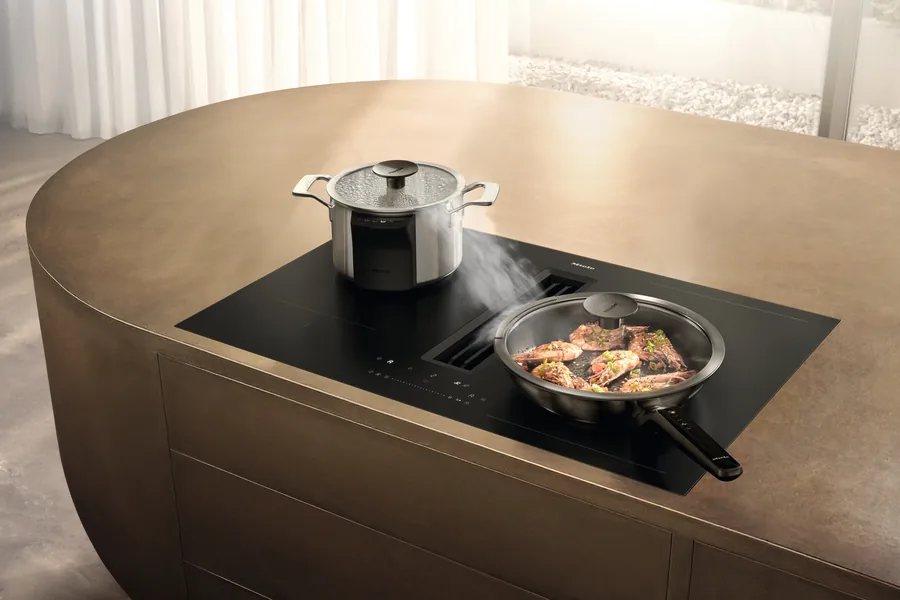 Placa de inducción Miele KMDA 8683 FL con extractor integrado en una isla de cocina color bronce, el vapor entra en la salida central